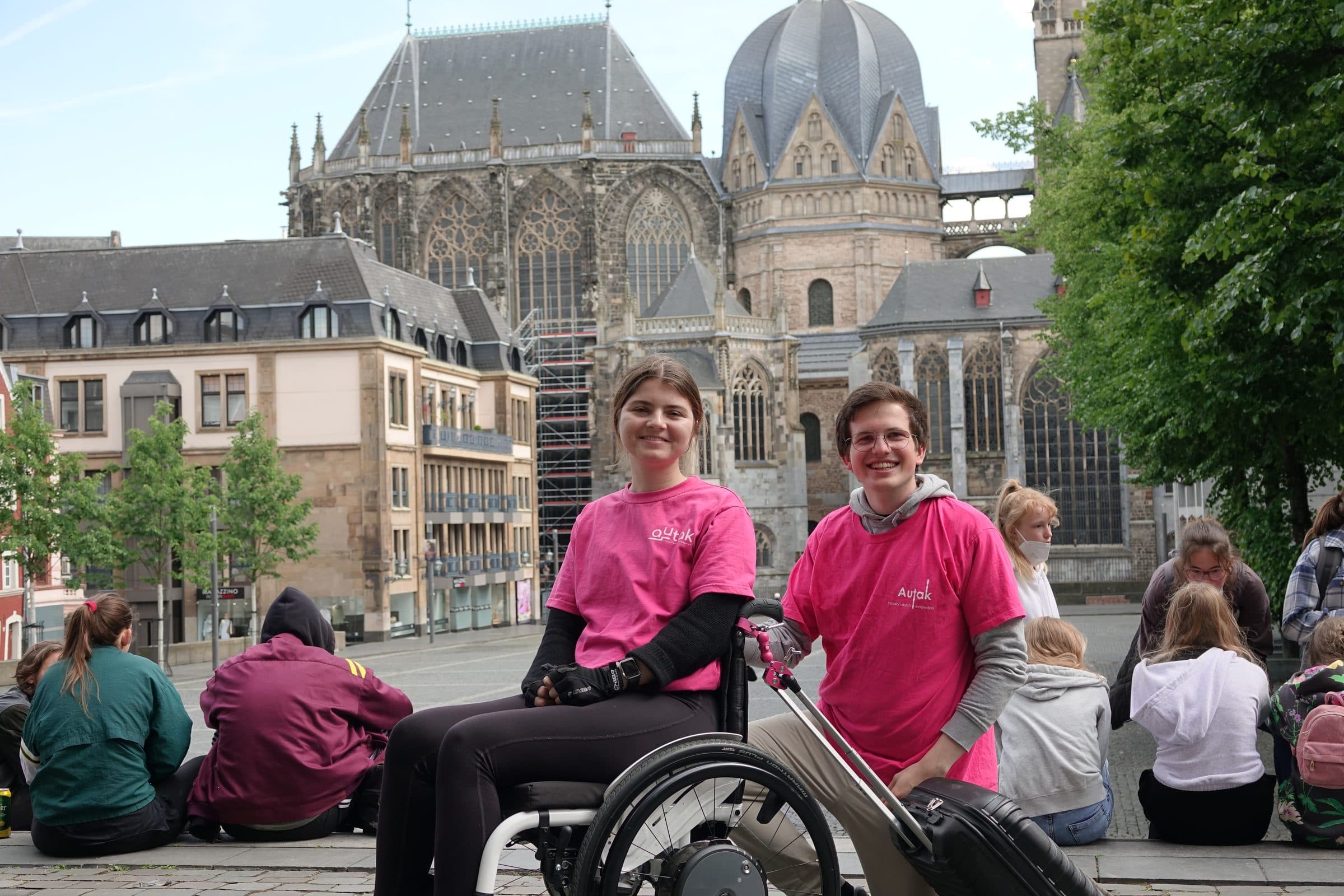 Zwei Mitglieder von Autak in pinken Vereins-T-Shirts lächeln neben einem Rollstuhl auf dem Katschhof vor dem Aachener Dom.