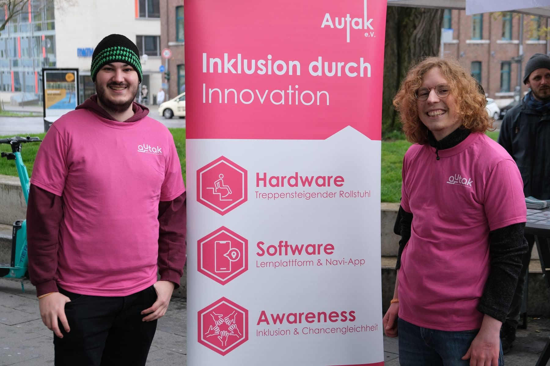 Zwei Autak-Mitglieder stehen neben einem Roll-Up mit der Aufschrift „Inklusion durch Innovation“ und den drei Säulen Hardware, Software und Awareness.